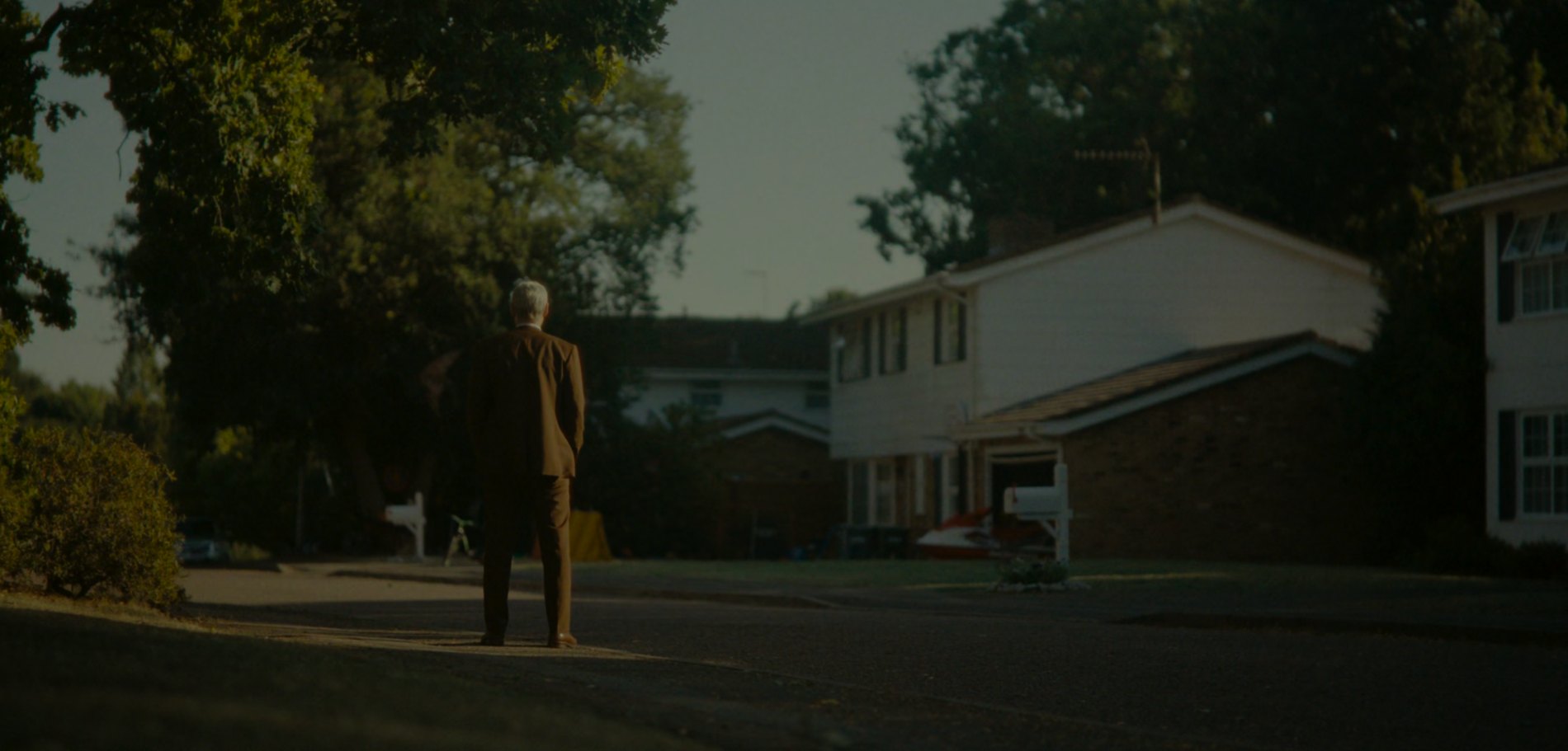 Mobius stands alone in a suburban street at golden hour, seen from behind, watching the houses of the ordinary life he chose.